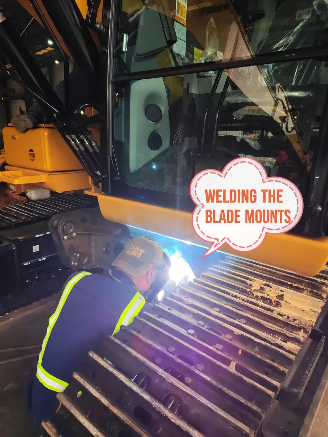 Technician welding a blade mounting bracket to the excavator lower-frame area.